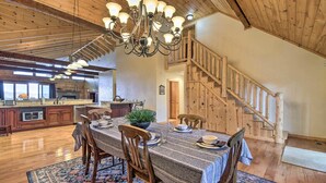 Un comedor espacioso con una gran mesa de madera, sillas y una araña. Una escalera con barandillas de madera conduce a un nivel superior.