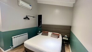 Habitación de hotel con cama, radiador, aire acondicionado, mesita de noche y otra mesita auxiliar.
