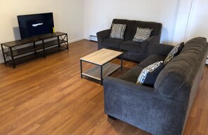 Ein Wohnzimmer mit einem dunkelgrauen Sofa, einem Holztisch und einem Fernseher auf einem schwarzen Standfuß.