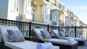 Fila de sillas de playa con almohadas y toallas estampadas, frente a un edificio con balcones y un cielo despejado.