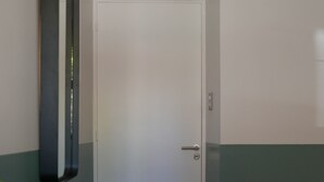 A room with a white door, a black vertical object on the wall, a white table, and a radiator. - Double Queen Size with Private Bathroom (Mendoza)