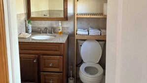 Ein kleines Badezimmer mit einem Holzkosmetikschrank, einem Waschbecken, einem Spiegel, einer Toilette und einem Regal mit Handtüchern.