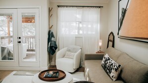 A cozy living room with a white armchair, a beige sofa, a small table with a tray, and a window with sheer curtains.