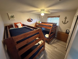 Una habitación con dos literas, un ventilador de techo, una ventana con persianas y una mesita de noche de madera.
