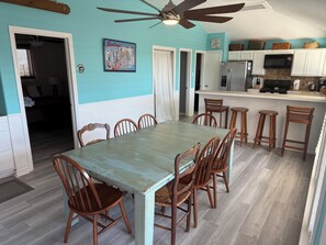 Un comedor con mesa y sillas de madera, ventilador de techo y una zona de cocina con refrigerador y microondas.