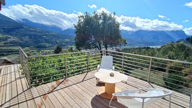 A wooden deck with a table and chairs, overlooking a mountainous landscape with vineyards. - Spacious apartment with magnificent view and terrace, on the 1st floor of a villa. (EMBRUN)