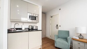 Une cuisine compacte équipée d’un four à micro-ondes, d’une bouilloire et d’une machine à café, ainsi qu’un fauteuil turquoise et une table de chevet avec une lampe.