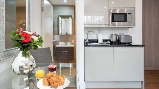 Une cuisine moderne équipée d’un four à micro-ondes, d’une bouilloire et d’un grille-pain. On y trouve également un coin repas avec une table, un vase rempli de fleurs, une assiette de croissants et un verre de jus d’orange.