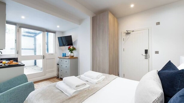 A hotel room with a bed, a desk with a flat-screen TV, a wardrobe, and a bathroom door. - PBC | Studio Apartment in Bayswater (LS2) (London)