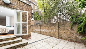 A brick house with a small backyard, a wooden fence, and a door leading to a room with a bed. - PBC | Large Studio w/Patio in Bayswater (LS4) (London)
