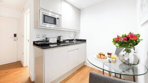 A compact kitchen with white cabinets, a microwave, and a coffee maker. There's a glass table with a vase of flowers and a plate of croissants. - PBC | Large Studio w/Patio in Bayswater (LS4) (London)