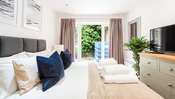 A modern hotel room with a bed, a TV, a desk, and a view of the garden. - PBC | Large Studio w/Patio in Bayswater (LS4) (London)