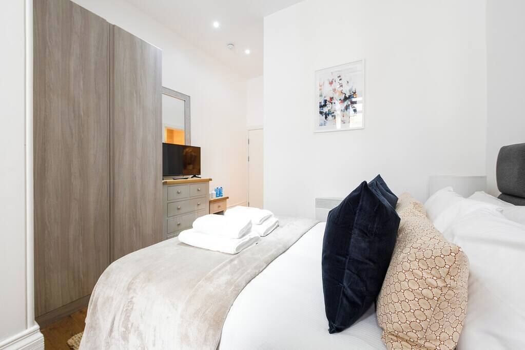 Ein modernes Schlafzimmer mit einem großen Bett, einem Kleiderschrank, einem Nachttisch mit Pflegeartikeln und einem gerahmten Kunstwerk an der Wand.