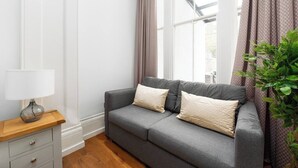 Ein graues Sofa mit zwei Kissen, ein Holznachttisch mit einer Lampe und ein Blumentopf.