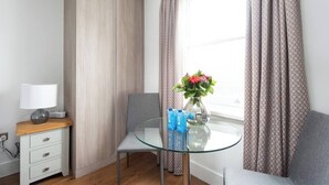 Una habitación pequeña con una mesa de vidrio, una silla, un sofá, una lámpara, un jarrón con flores y cortinas.