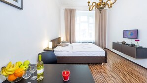 A hotel room with a bed, bedside table with fruit, wine, and candle, a TV on a wall, and a chandelier. - Central & Parking Assistance (Prague)