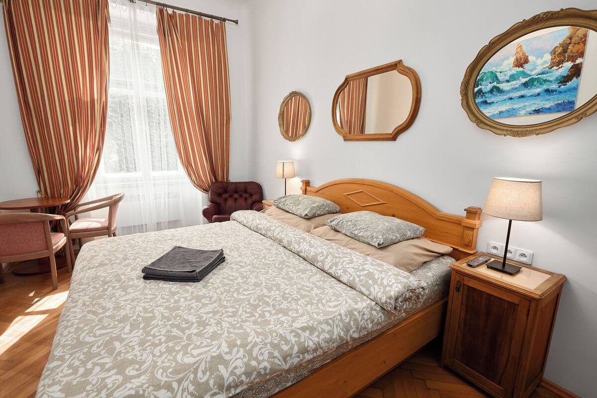 A neatly made bed with a patterned bedspread, a bedside table with a lamp, a chair, and a painting of a seascape on the wall.