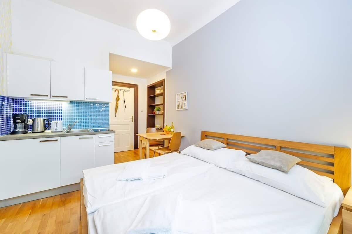 A bedroom with a large bed, a wooden headboard, and a bedside table. There is a kitchenette with a stove, a sink, and a countertop. A dining area with a table and chairs is visible in the background.