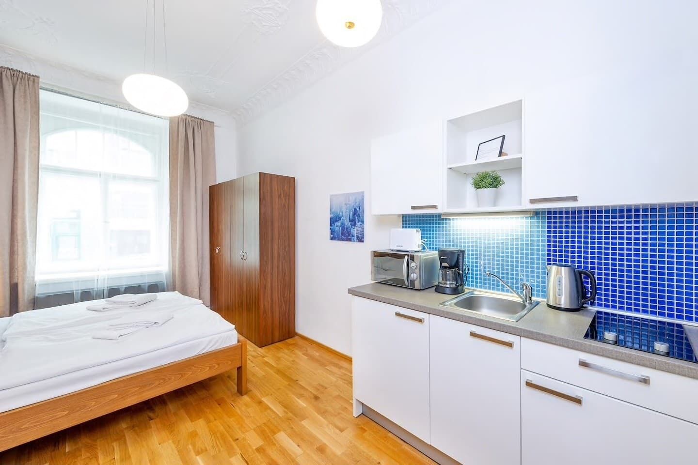 A modern hotel room with a wooden bed, a kitchenette with a blue backsplash, and a large window with curtains.