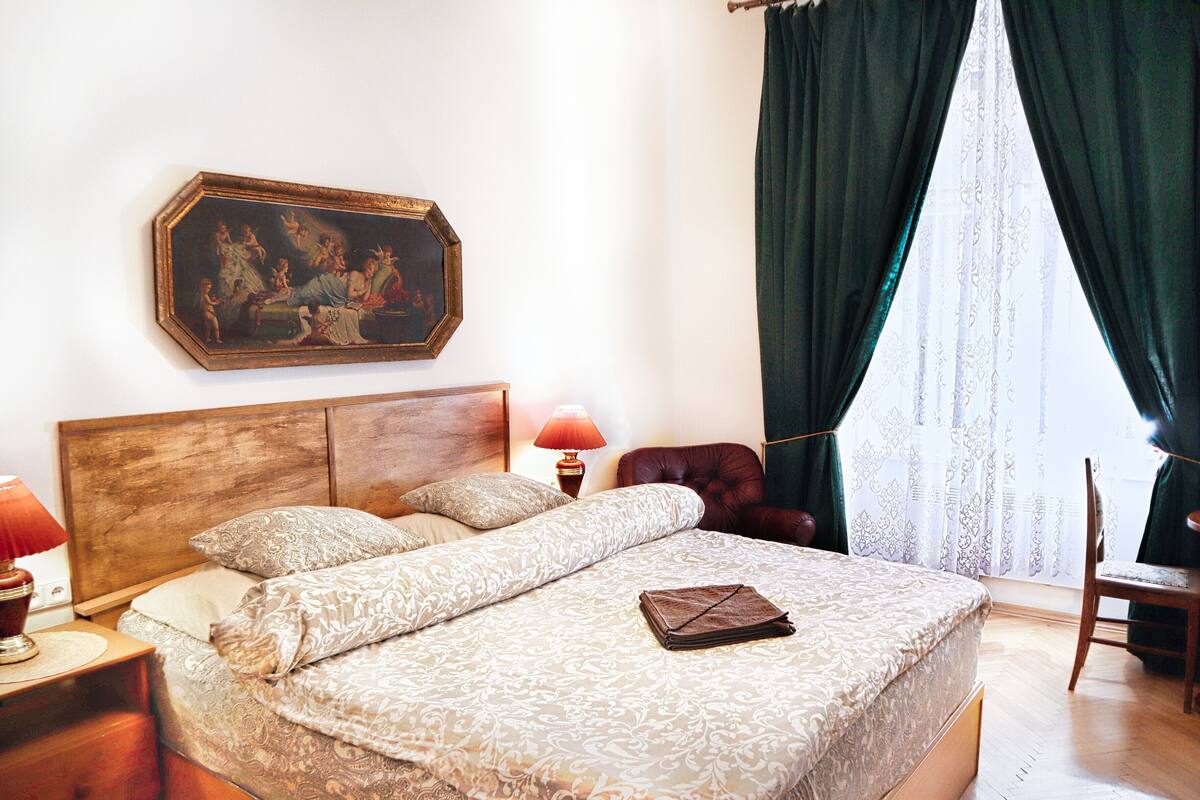 A bedroom with a wooden headboard, a bed with patterned bedding, a nightstand with a lamp, a chair, and a painting on the wall.