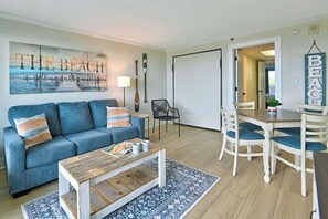 Un salon avec un canapé bleu, une table basse, une table à manger avec des chaises, un mur orné d’une œuvre d’art sur le thème de la plage et une porte donnant sur une autre pièce.