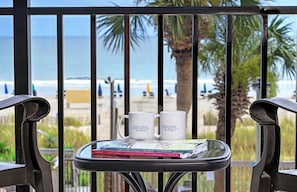 A balcony with two white mugs labeled 'Coastal Real Estate' and a magazine on a glass table, overlooking a beach with palm trees and the ocean.