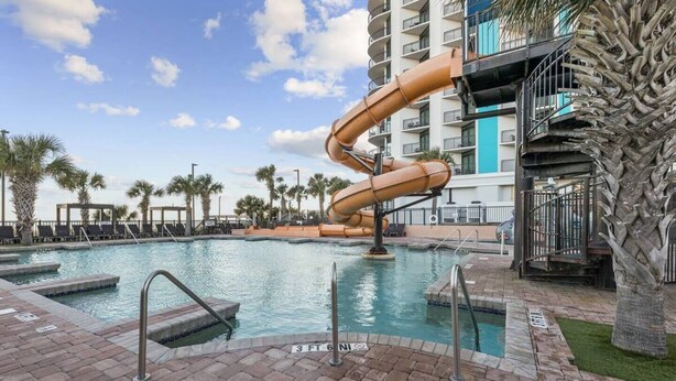 A pool area with a water slide, palm trees, and a multi-story building in the background.