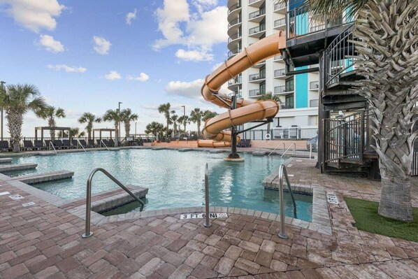 A pool area with a water slide, palm trees, and a multi-story building in the background.