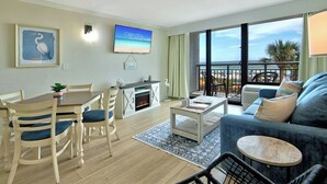 A living room with a sofa, coffee table, and dining area with chairs. There's a TV mounted on the wall and a balcony with a view.