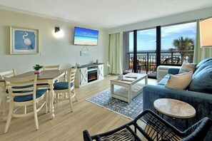 A living room with a sofa, coffee table, and dining area with chairs. There's a TV mounted on the wall and a balcony with a view.