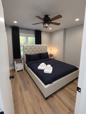A modern bedroom with a tufted headboard, a ceiling fan, a bedside table with a lamp, and a window with curtains. - 6 bedrooms! Pool Table, Gaming Area, Firepit & More (Joliet)