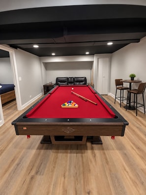 A pool table with a red felt surface, surrounded by wooden flooring, in a room with a dark ceiling and white walls. - 6 bedrooms! Pool Table, Gaming Area, Firepit & More (Joliet)