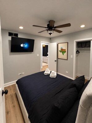 Un dormitorio con cama, ventilador de techo, televisor montado en la pared y puerta que da a otra habitación.