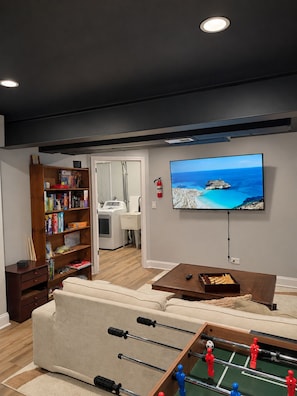 A living room with a sofa, a foosball table, a television mounted on the wall, and a bookshelf.