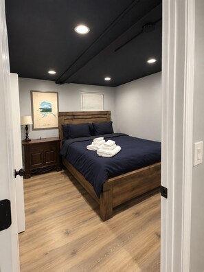 Un dormitorio con cama de madera, mesita de noche, mapa enmarco y luces empotradas en el techo.