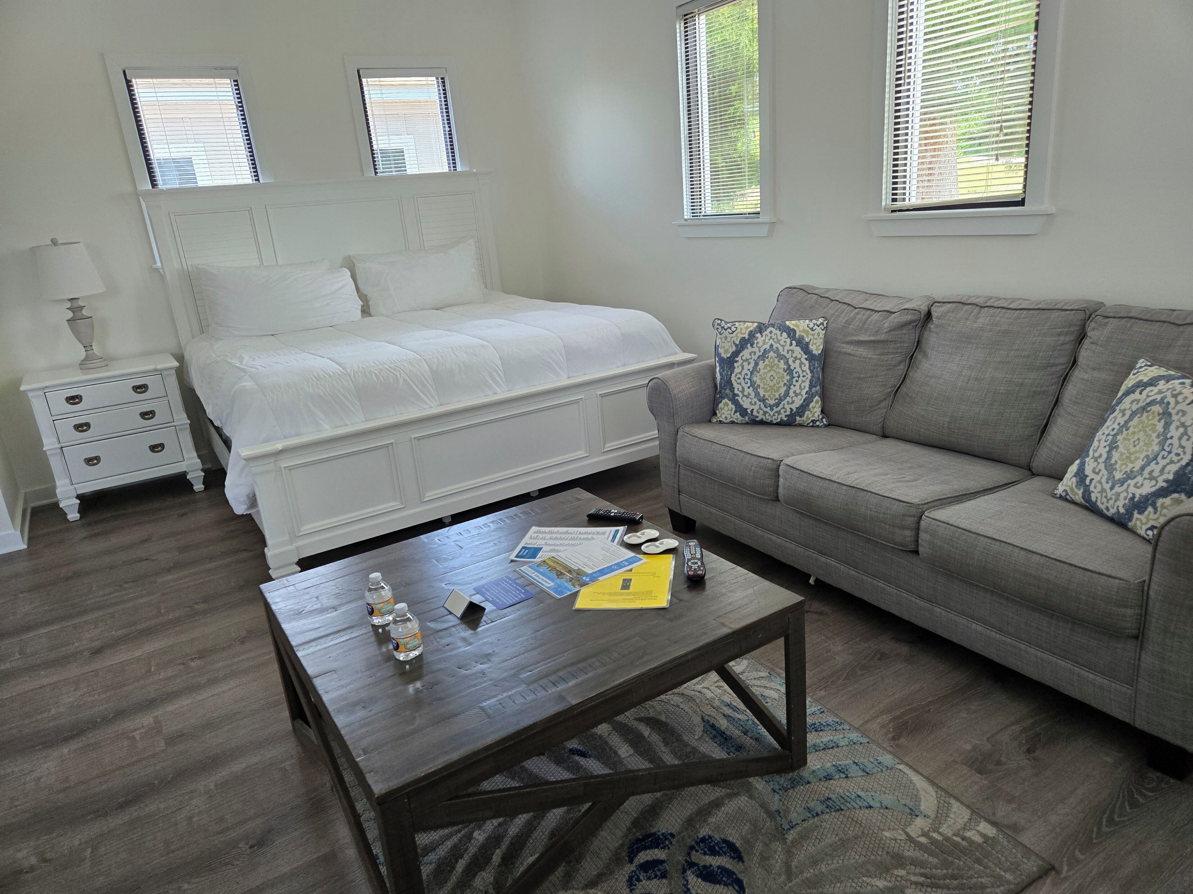 A bedroom with a bed, a grey sofa, a wooden coffee table, a nightstand, and two windows with blinds.