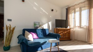 Een woonkamer met een blauwe bank, een klein rond tafeltje, een televisie op een houten kast en een raam met gordijnen.