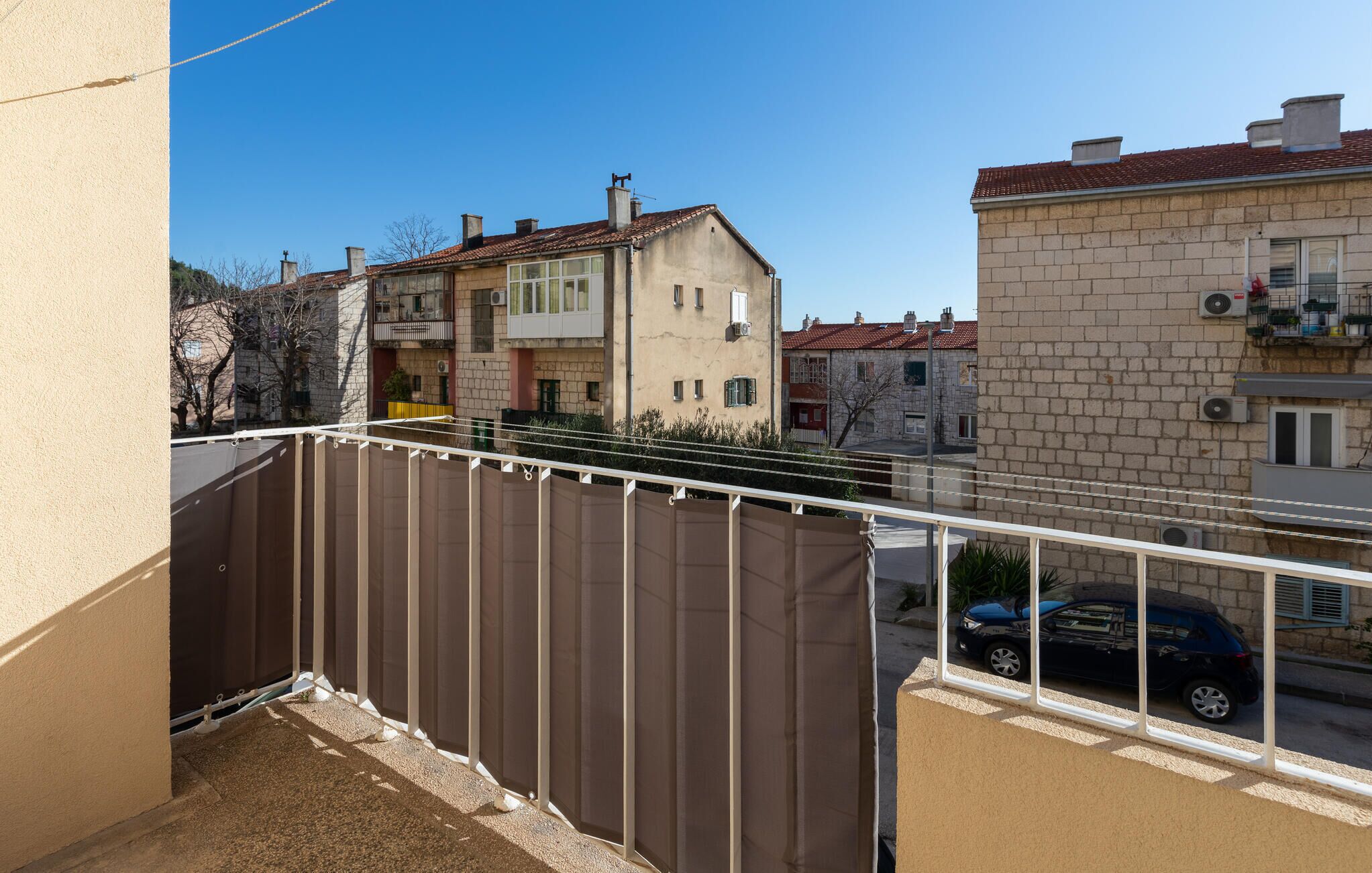Ein Balkon mit brauner Metallreling, einer beigen Wand und Blick auf benachbarte Gebäude mit roten Ziegeldächern.