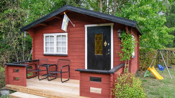 Une petite cabane en bois avec une porte noire, une fenêtre et une véranda dotée de chaises en métal.