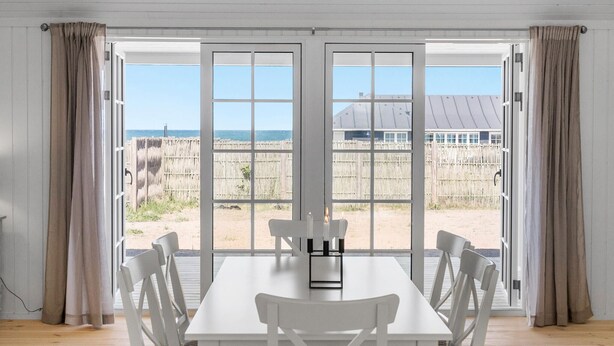 Een kamer met een grote glazen schuindeur die uitkomt op een uitzicht op het strand, een witte tafel en vier stoelen.