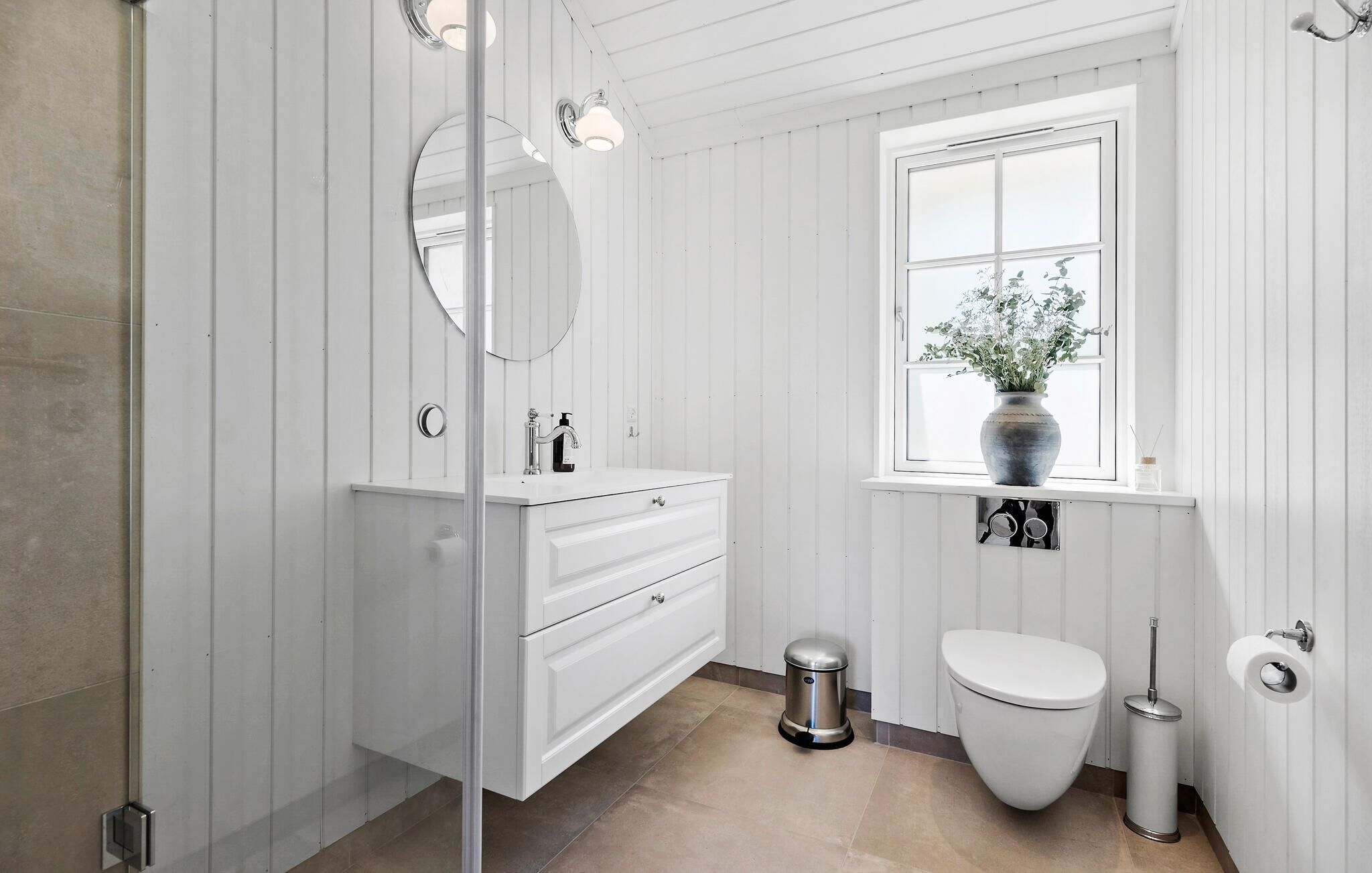 A bathroom with white wooden paneling, a round mirror, a white sink with a faucet, a toilet, and a window with a vase of flowers.