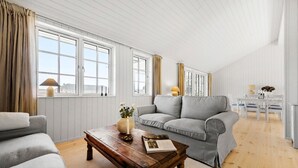 Een ruime woonkamer met een grijze bank, een houten salontafel, en een eethoek met een witte tafel en stoelen.