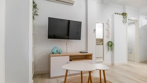 Un saló modern amb un televisor de pantalla plana fixat a la paret, un moble de fusta amb objectes decoratius, una taula rodona petita amb dues cadires, i una catifa teixida al terra.