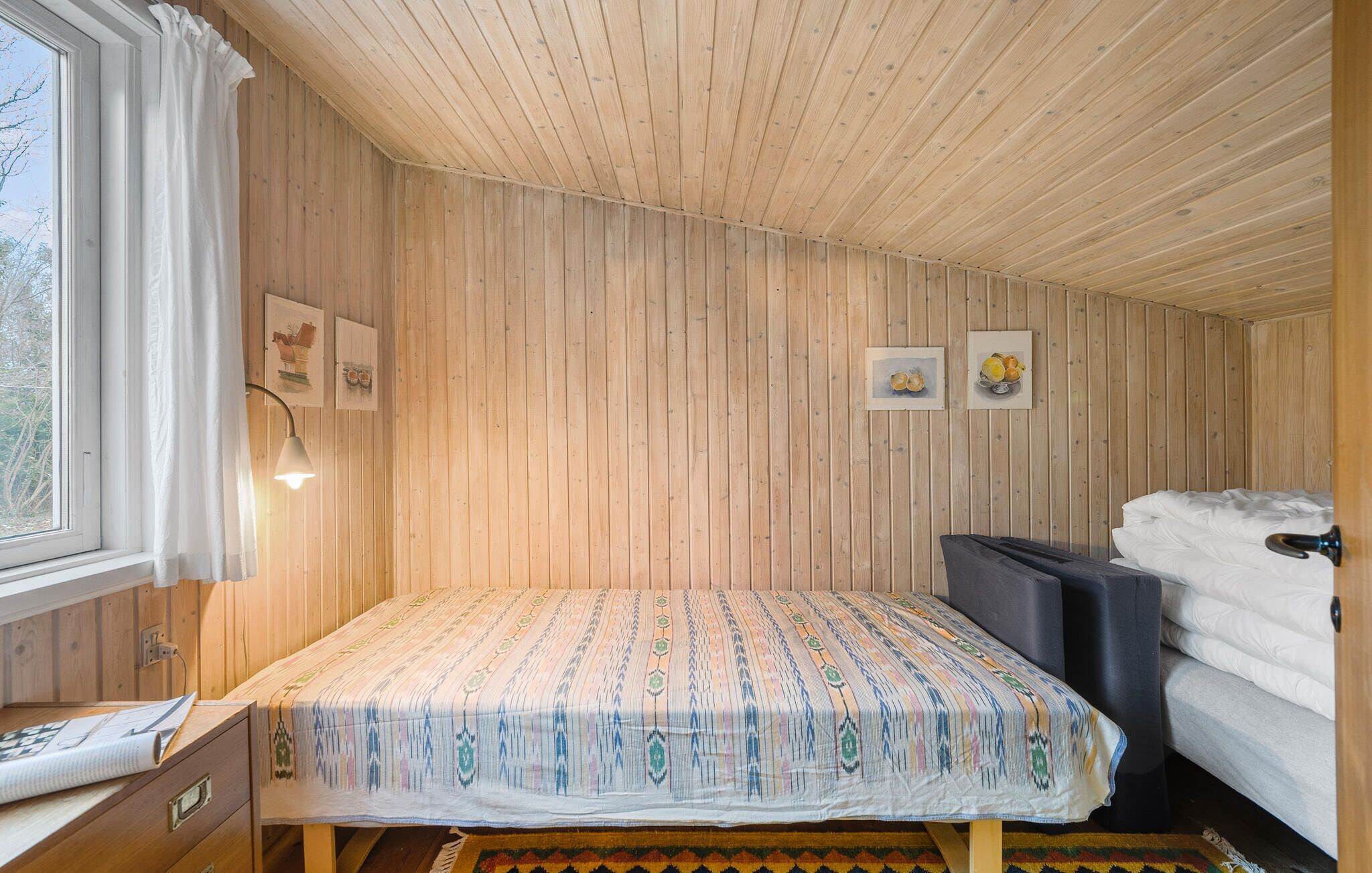 Ein kleiner, gemütlicher Raum mit Holzwänden und geneigter Decke. Er verfügt über ein Bett mit gemustertem Überwurf, einen Nachttisch mit Lampe sowie ein Fenster mit Vorhängen.