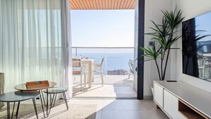 Un salon moderne doté d’un grand téléviseur, d’un coin repas avec une table et des chaises, et d’un balcon offrant une vue sur la mer.