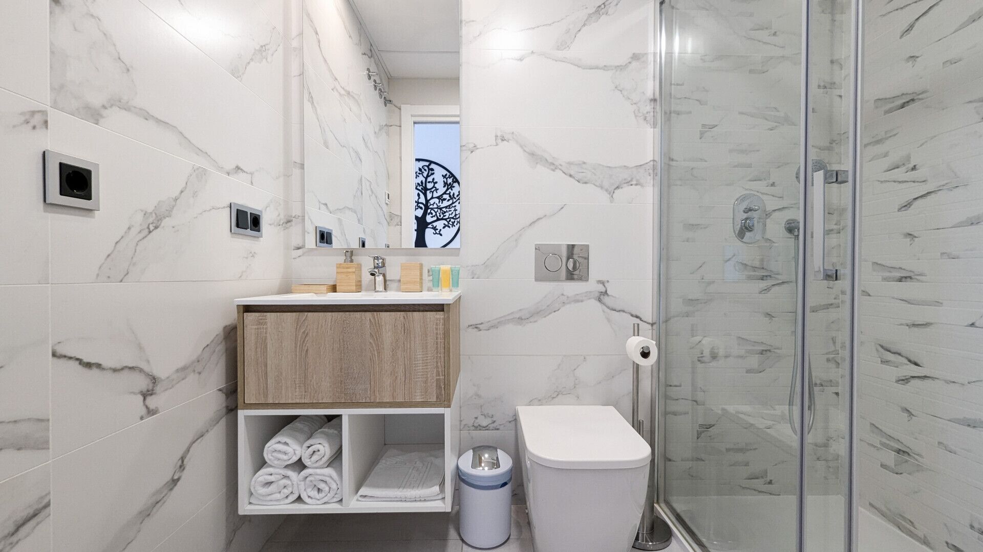 Un baño moderno con paredes de mármol, un tocador de madera, cabina de ducha de vidrio y un inodoro.