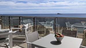 A balcony with a view of the sea, furnished with white chairs and a table with a bowl of fruit.
