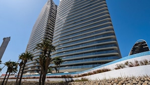 Two tall, modern buildings with horizontal lines of windows, surrounded by palm trees and a landscaped area with white and blue elements.
