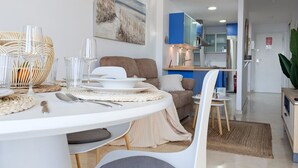 Un comedor moderno con una mesa blanca dispuesta para cuatro comensales, un sofá y una cocina al fondo.