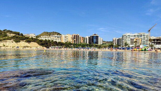 Un paisaje urbano costero con aguas transparentes que reflejan edificios, una playa arenosa y un fondo de colinas.